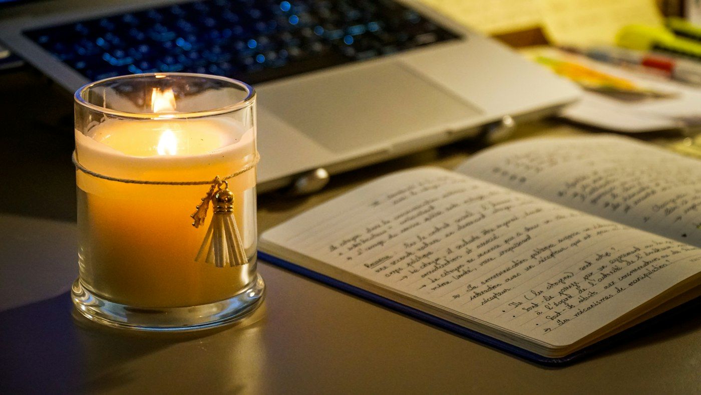 Brennende Kerze neben aufgeschlagenem Notizbuch und Laptop